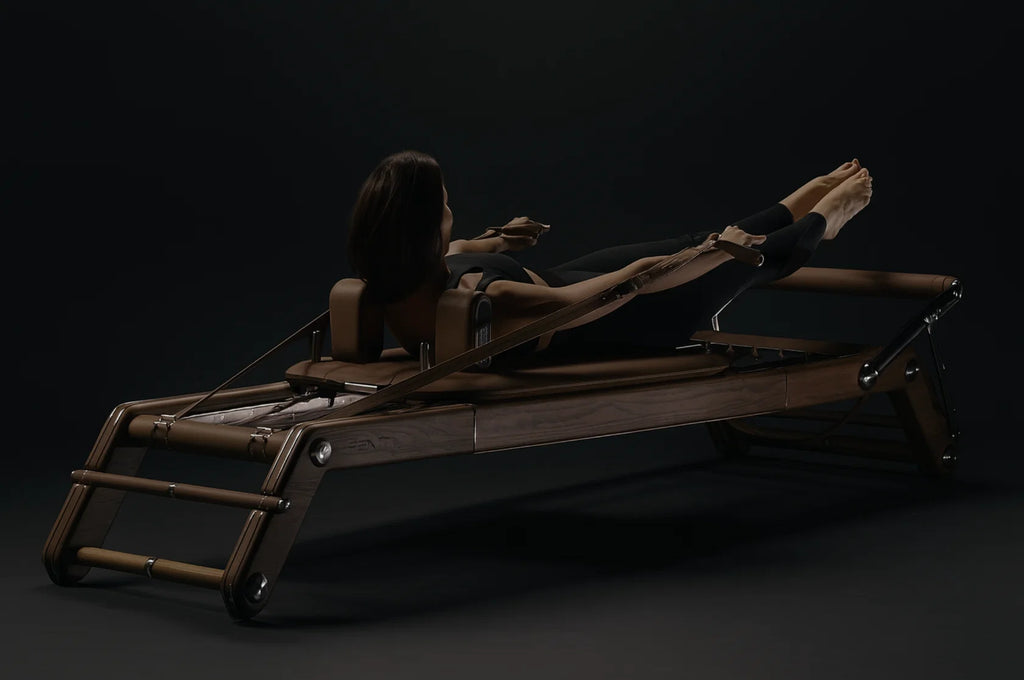 Person in black exercising on a dark wood Pilates reformer against a near-black background