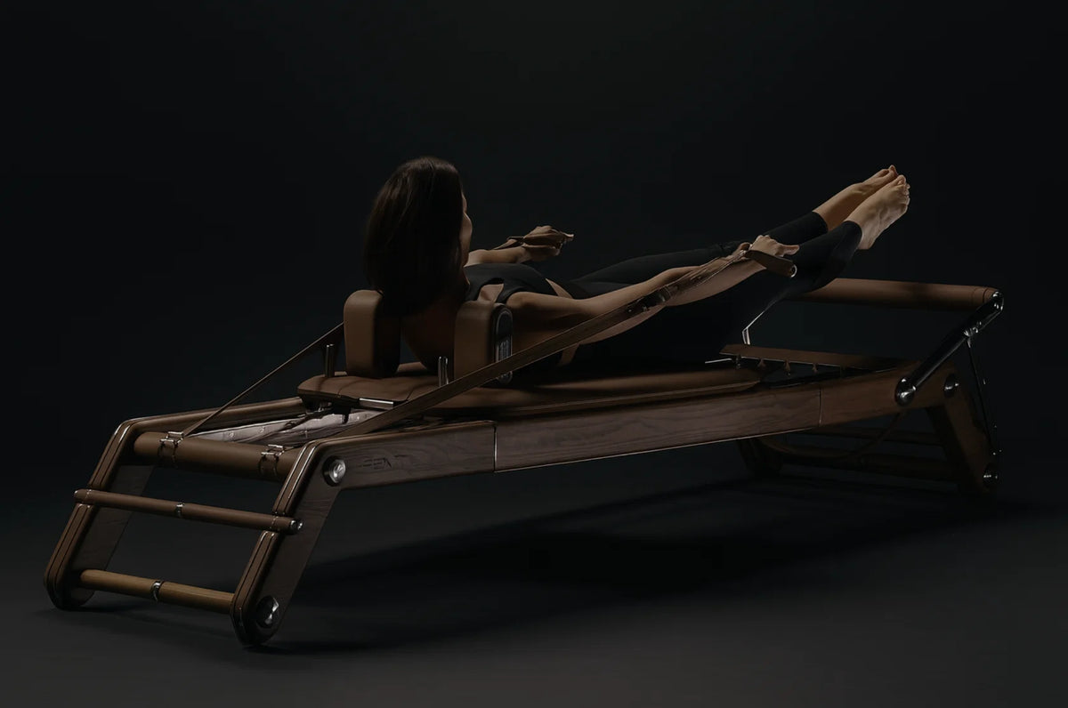 Person in black exercising on a dark wood Pilates reformer against a near-black background