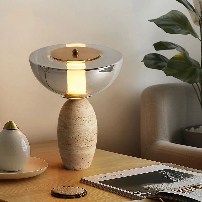 Silver bowl-shade table lamp with textured base on a wood table beside books and a chair
